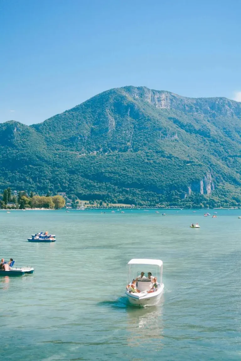Clear blue waters of Lake Annecy with boats in the foreground and forested mountains in the background